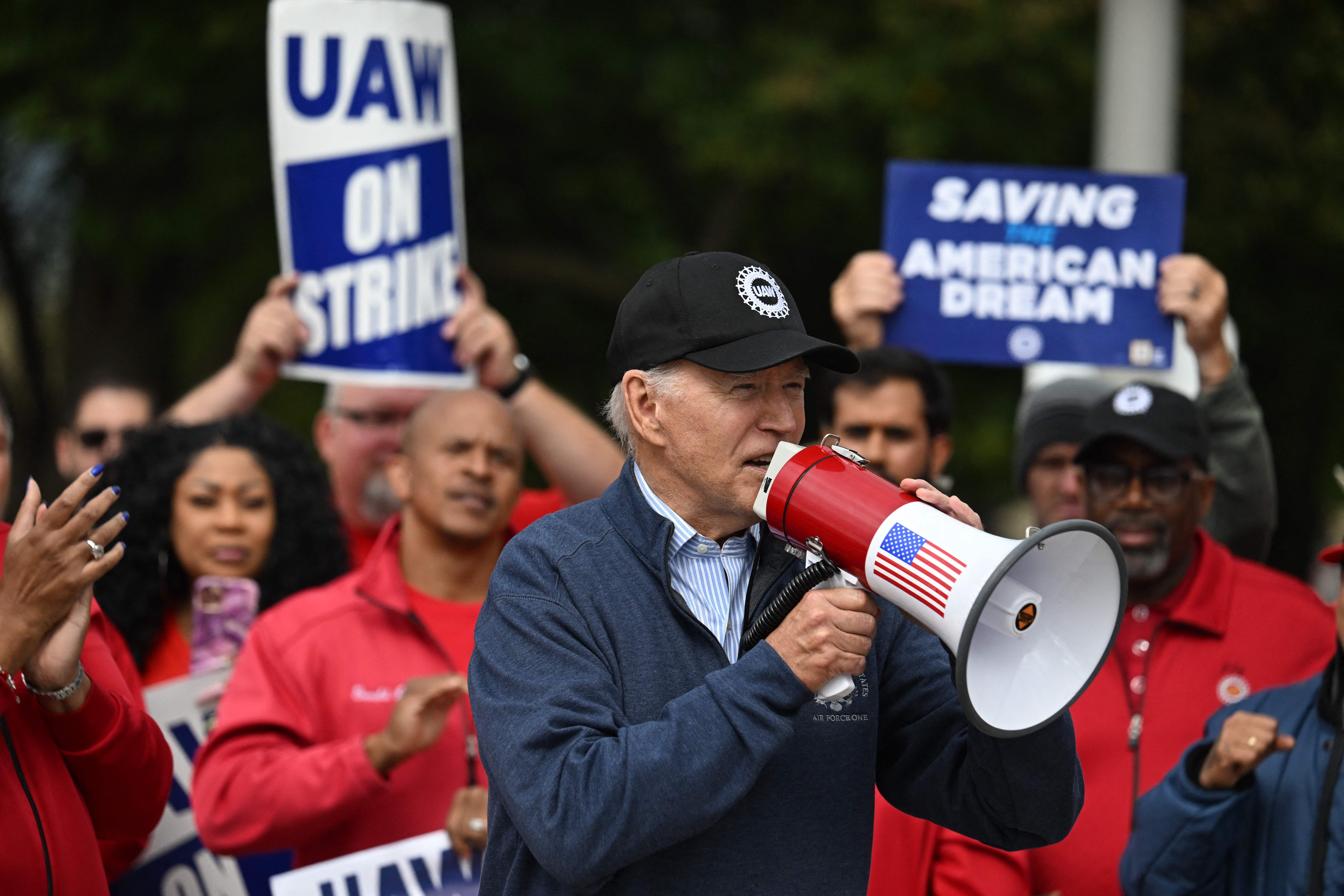 Biden speaking into a bullhorn while standing in front of striking auto workers holding signs saying "UAW on Strike" and "Saving the American Dream"